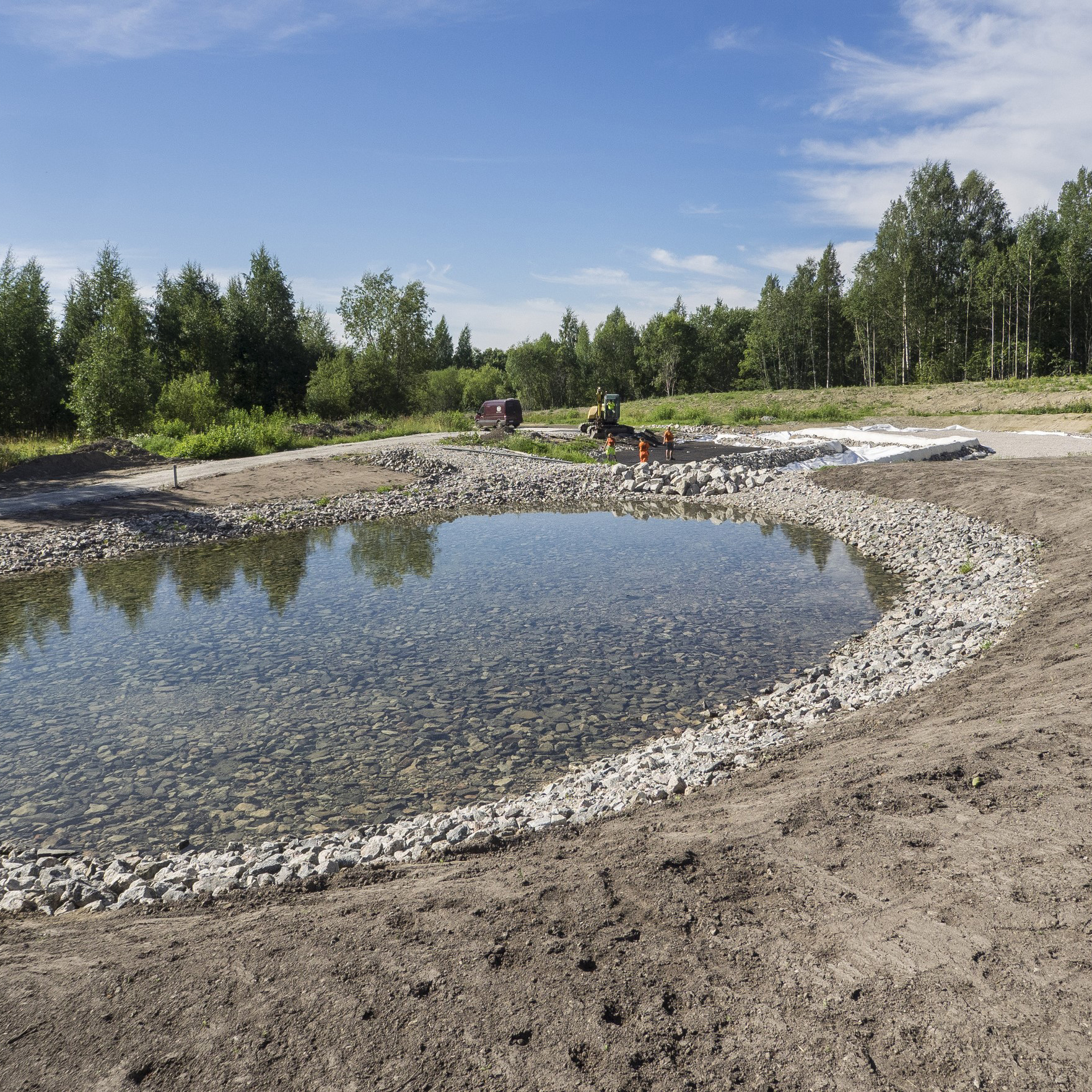 Nyanlagd dagvattendamm med stenfilter och arbetsmaskiner under byggskede i naturlig omgivning.
