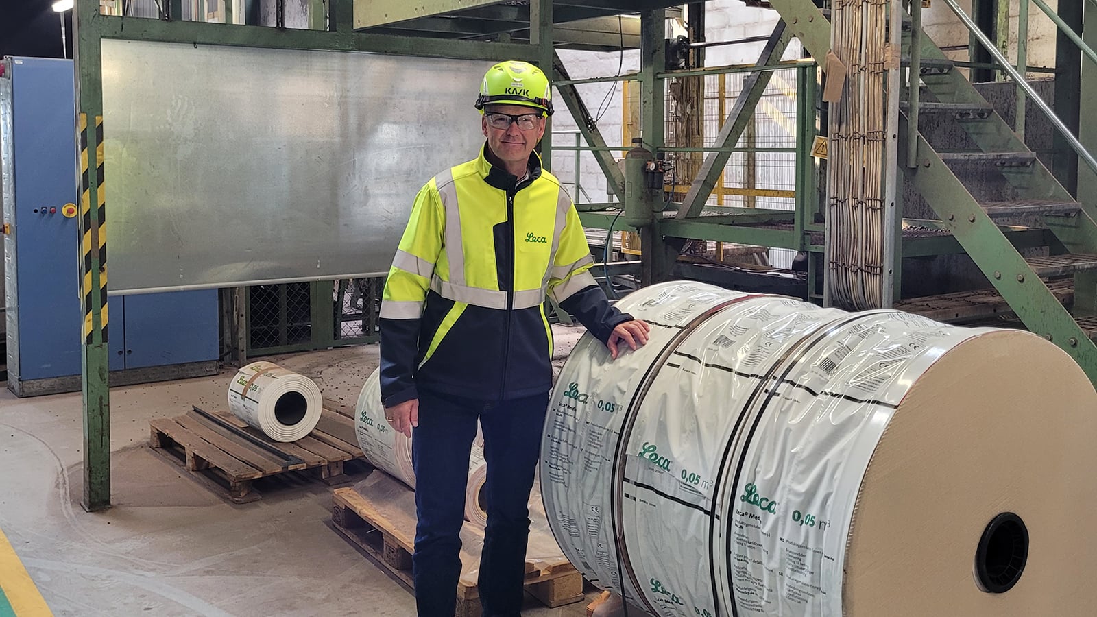 Man i gul reflexjacka och hjälm med Leca-logotyp står i en industrimiljö bredvid stora rullar med förpackningsmaterial.