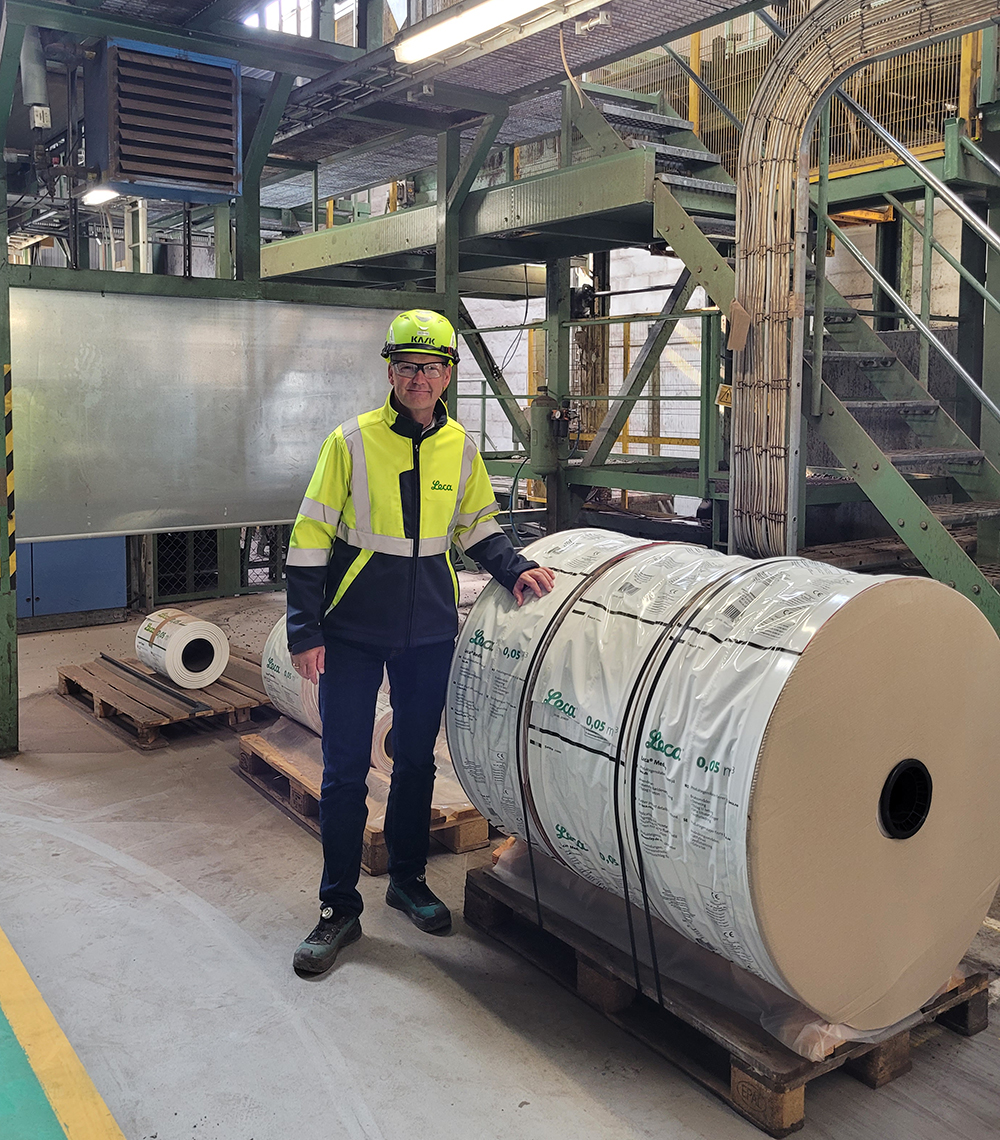 Man i gul reflexjacka och hjälm med Leca-logotyp står i en industrimiljö bredvid stora rullar med förpackningsmaterial.