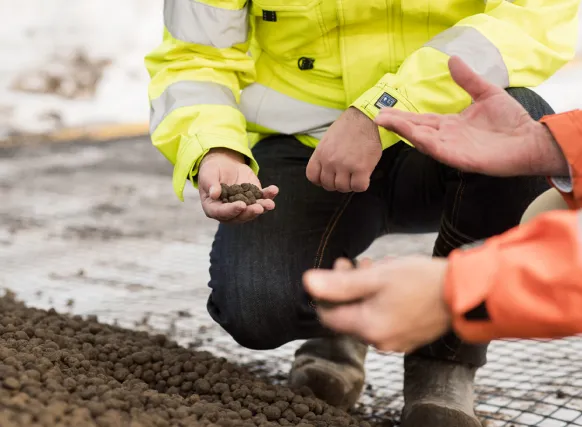 Två personer i arbetskläder undersöker Leca lättklinker vid en anläggningsyta, en håller kulor i handen.