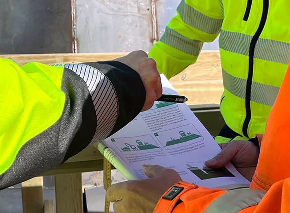närbild på en person med varseljacka som håller i en teknisk handbok, två andra personer står runtomkring och en av dom pekar på sidan med en penna