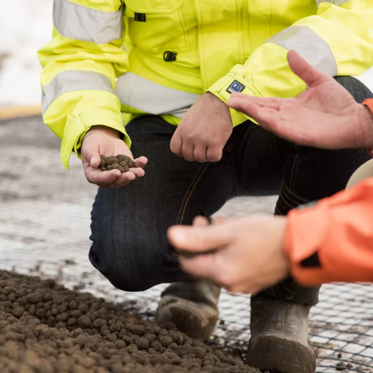 två personer i byggarbetskläder sitter på hur och gestikulrear med händerna och lyfter upp lecakulor från marken
