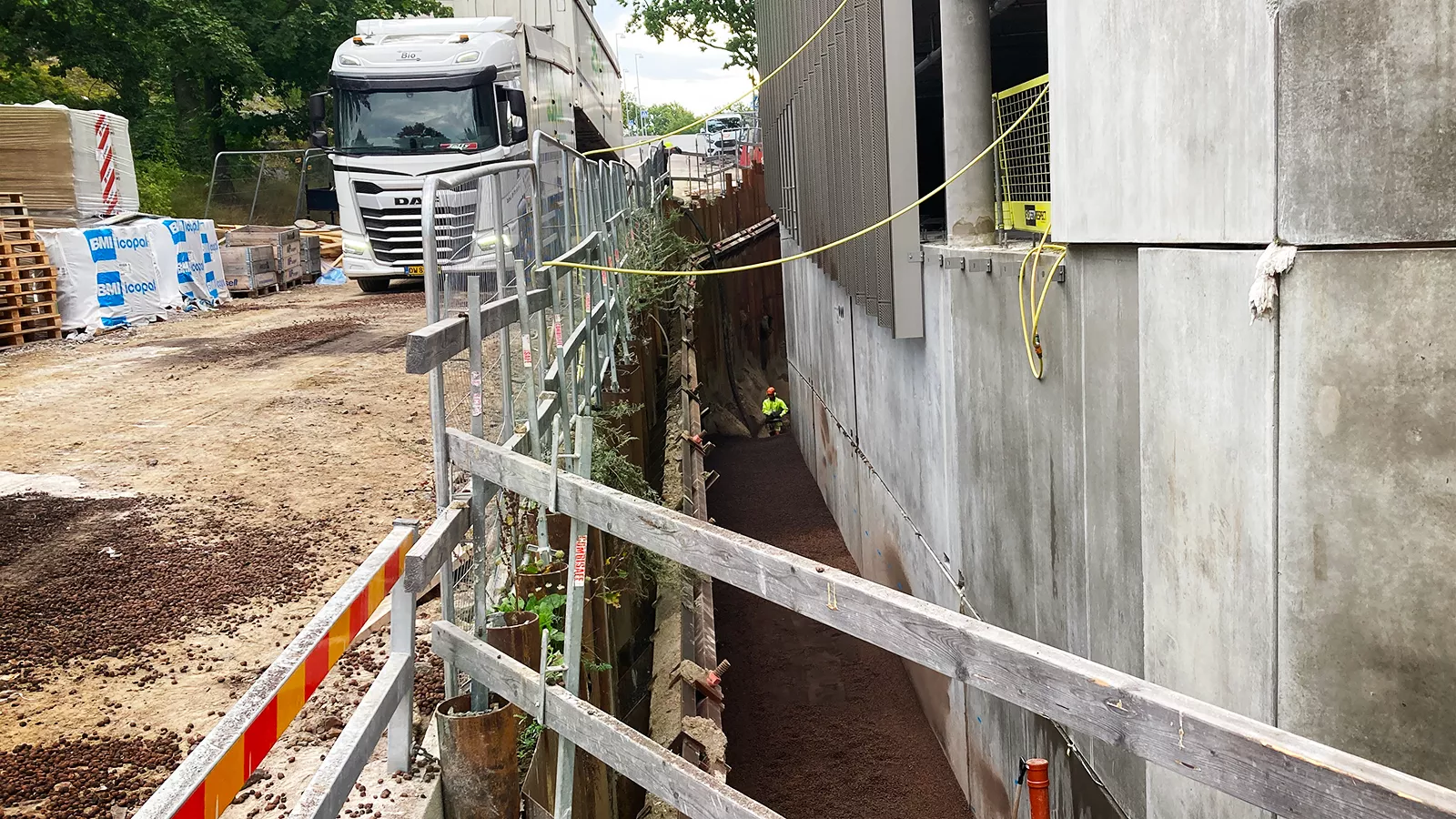 Smal schakt intill byggnadens fasad, delvis fylld med lättklinker, med arbetsställning och skyddsräcke längs kanten. En lastbil syns i bakgrunden.