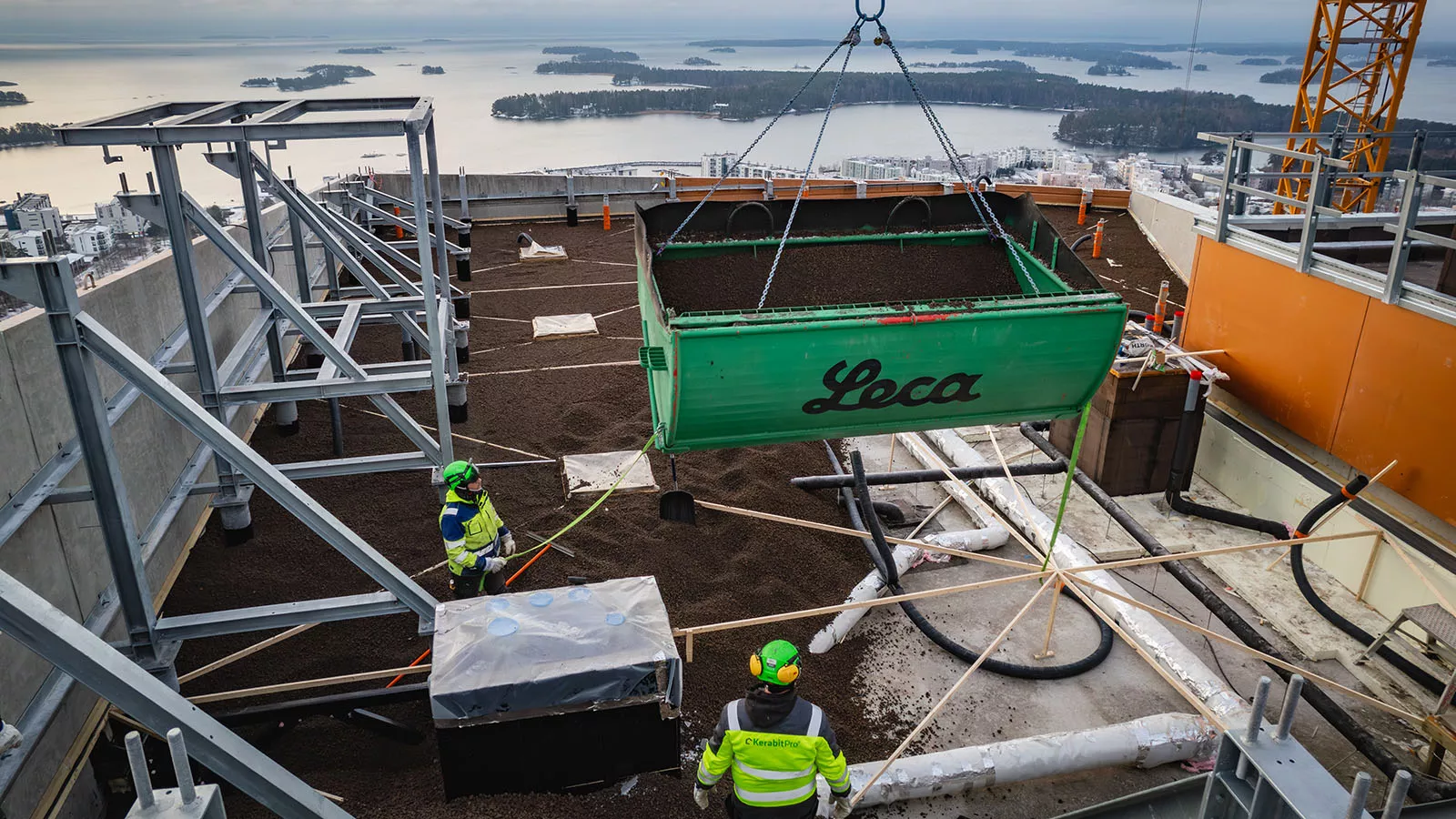 Arbetsplats på ett tak, där två byggarbetare i skyddsutrustning övervakar en grön container märkt "Leca" som lyfts med en kran.