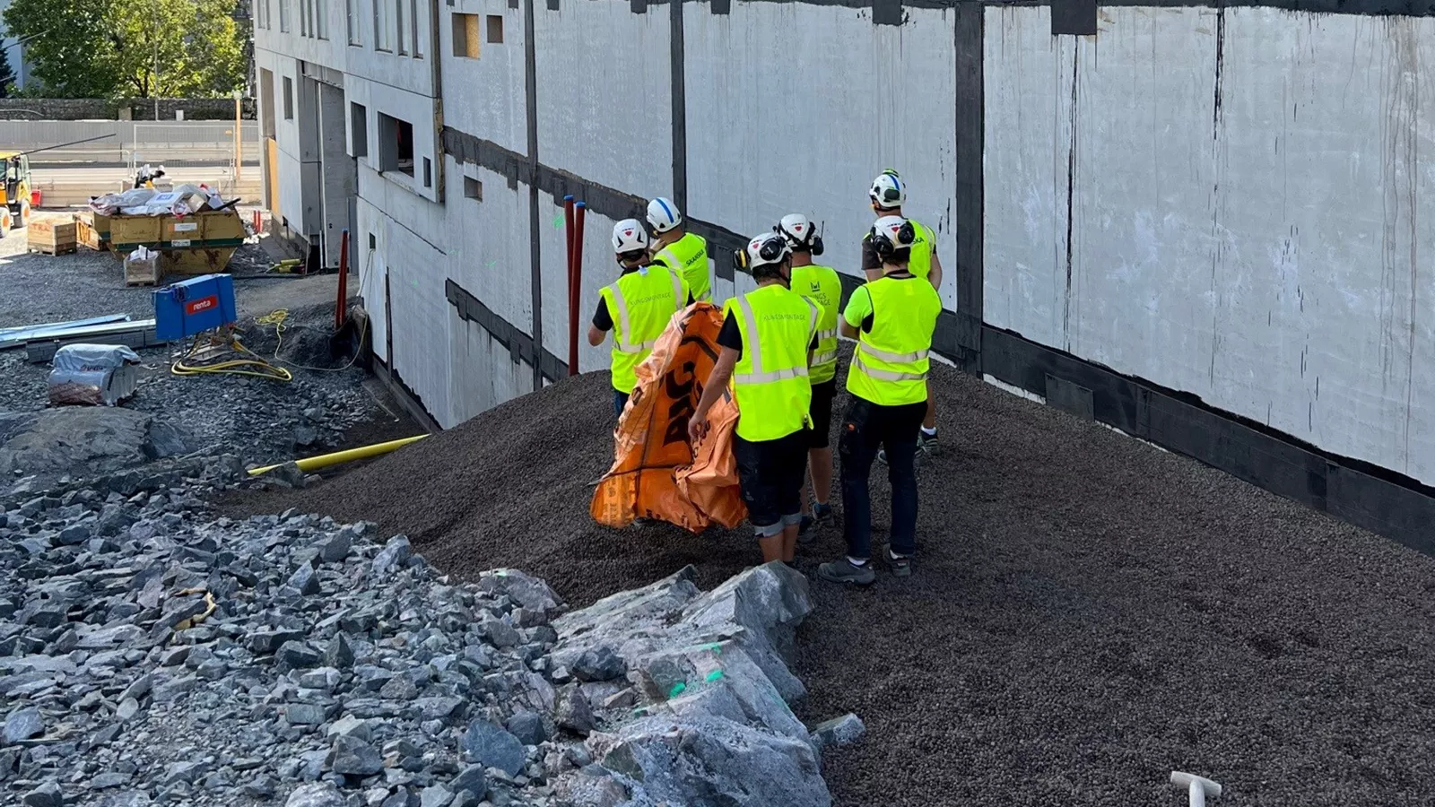 Sex personer i byggvästar står vid en husfasad, de står ovanpå en nylagd fyllning av Leca lättklinker som fyllt upp utrymmet mellan berg och husväggen