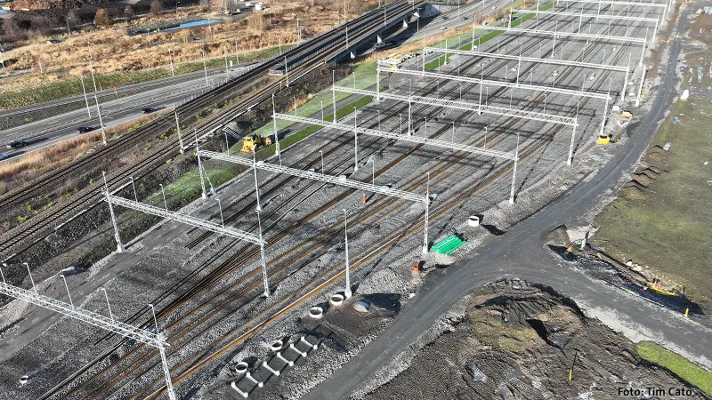 Flygfoto över pågående byggnation av Lärje uppställningsspår med flera parallella spår, kontaktledningsstolpar och grusballast, intill vägar och markarbeten i ett öppet landskap.