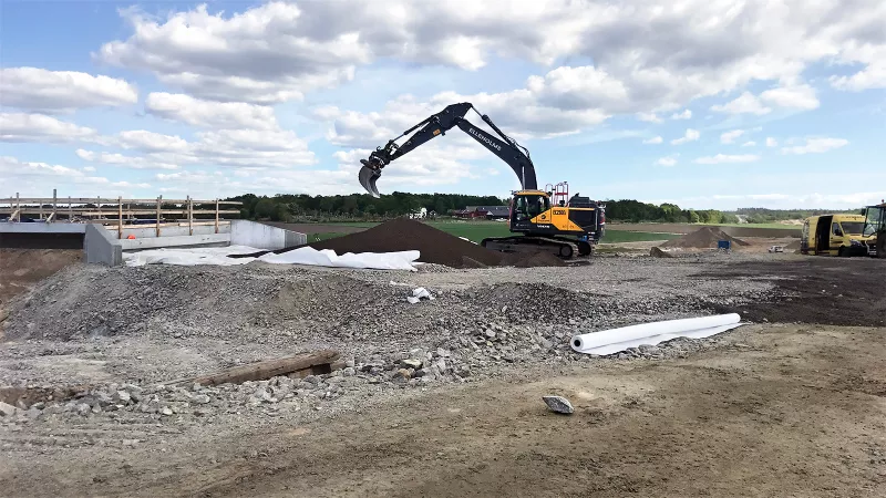 En grävmaskin arbetar med att flytta Leca Lättklinker vid en byggarbetsplats. På marken ligger geotextilduk och olika materialhögar. I bakgrunden syns ett lantligt landskap med träd och byggnader samt en klar himmel med moln.