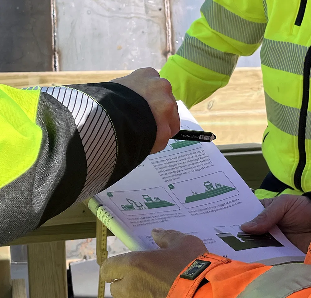 Två personer i varselkläder pekar och diskuterar ritning eller instruktion med illustrationer på en byggarbetsplats.