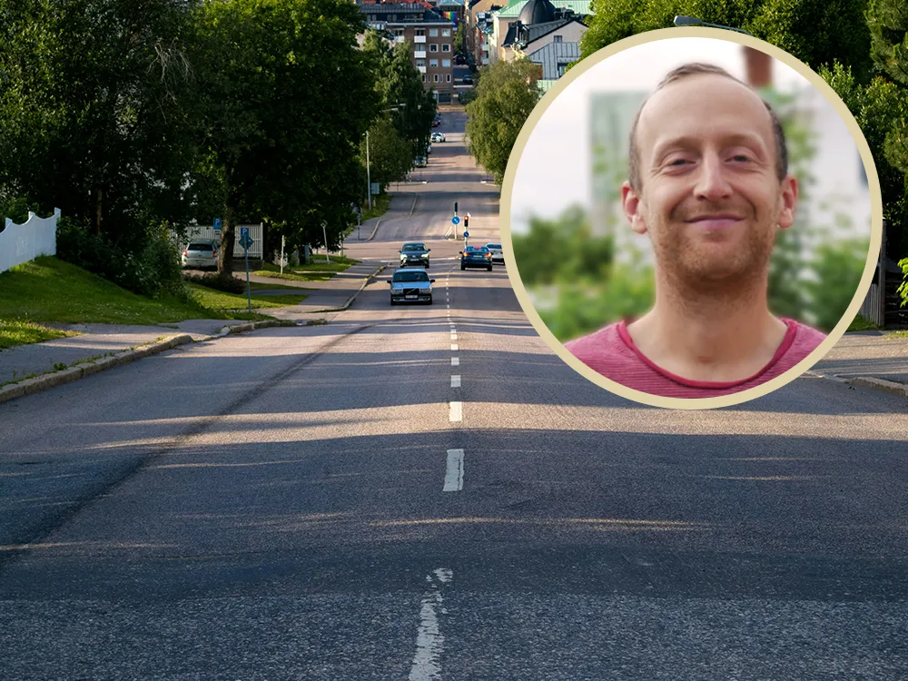 En bred stadsgata kantad av träd, med några bilar som kör i båda riktningarna. I förgrunden till höger syns ett infällt porträtt av en leende man med kort hår och röd t-shirt, mot en suddig grön bakgrund.