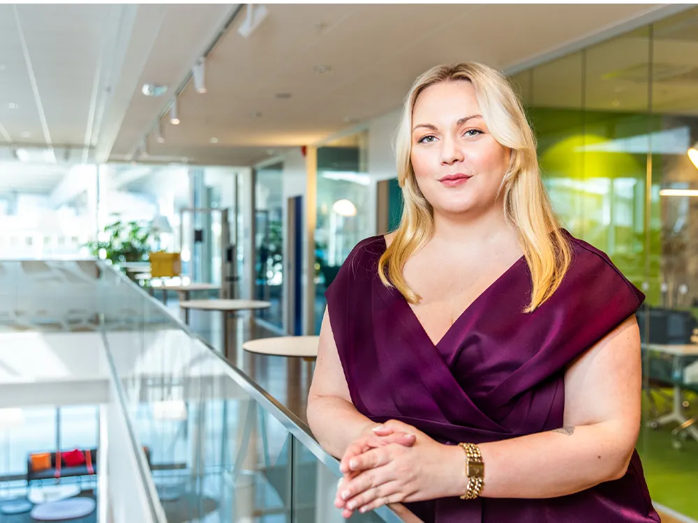 Amanda Borneke står lutad mot ett räcke i en modern kontorsmiljö, iklädd vinröd klänning och med händerna lätt sammanförda.