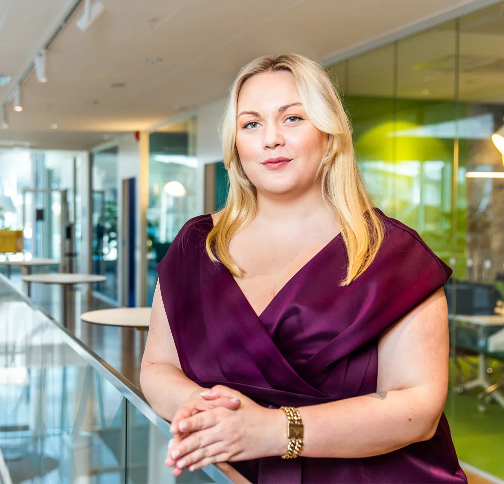 Amanda Borneke står lutad mot ett räcke i en modern kontorsmiljö, iklädd vinröd klänning och med händerna lätt sammanförda.