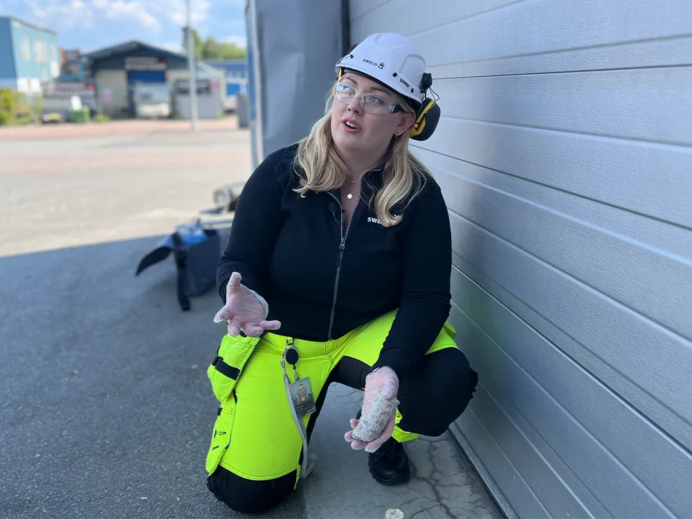 Kvinna i skyddshjälm och varselbyxor sitter på huk utomhus, håller i ett materialprov och pratar.