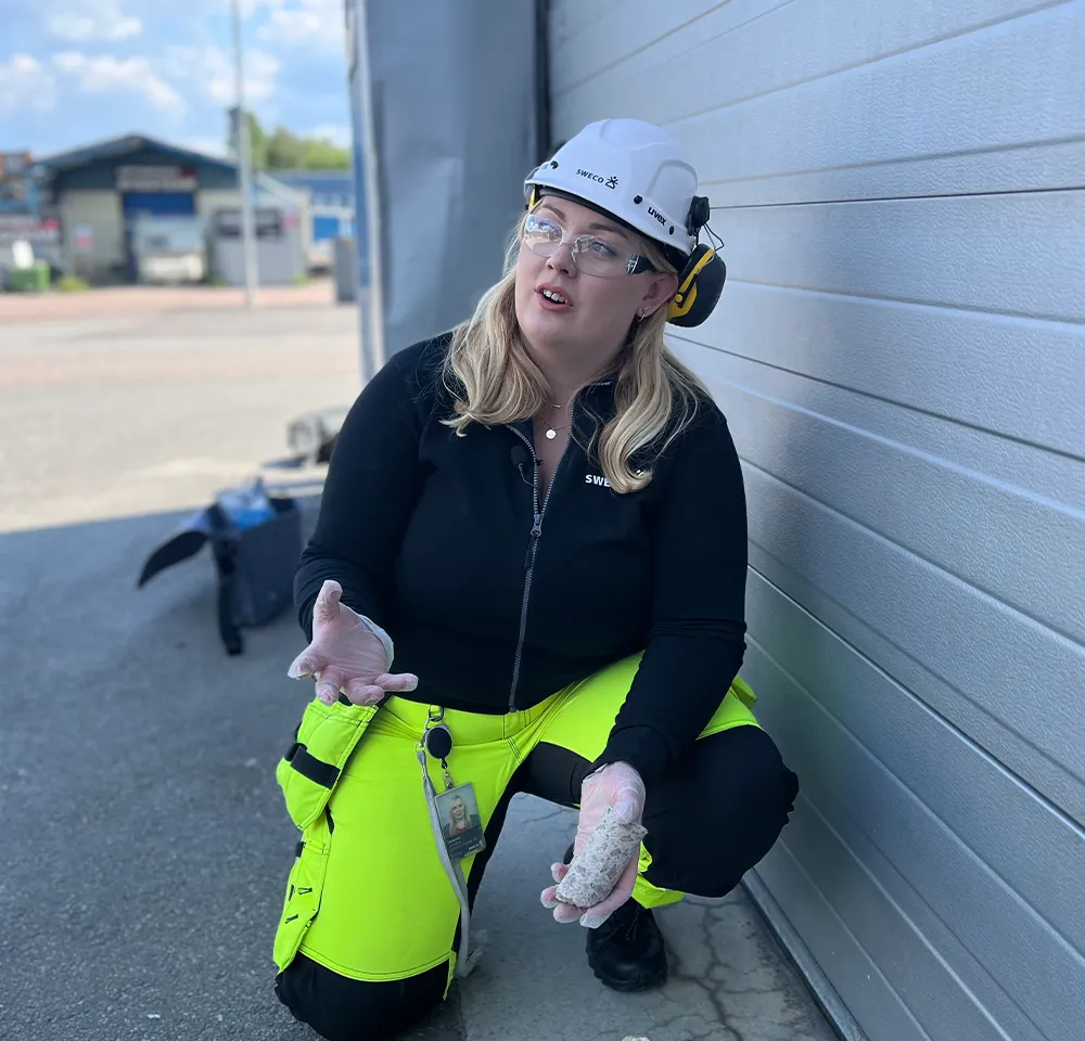 Kvinna i skyddshjälm och varselbyxor sitter på huk utomhus, håller i ett materialprov och pratar.