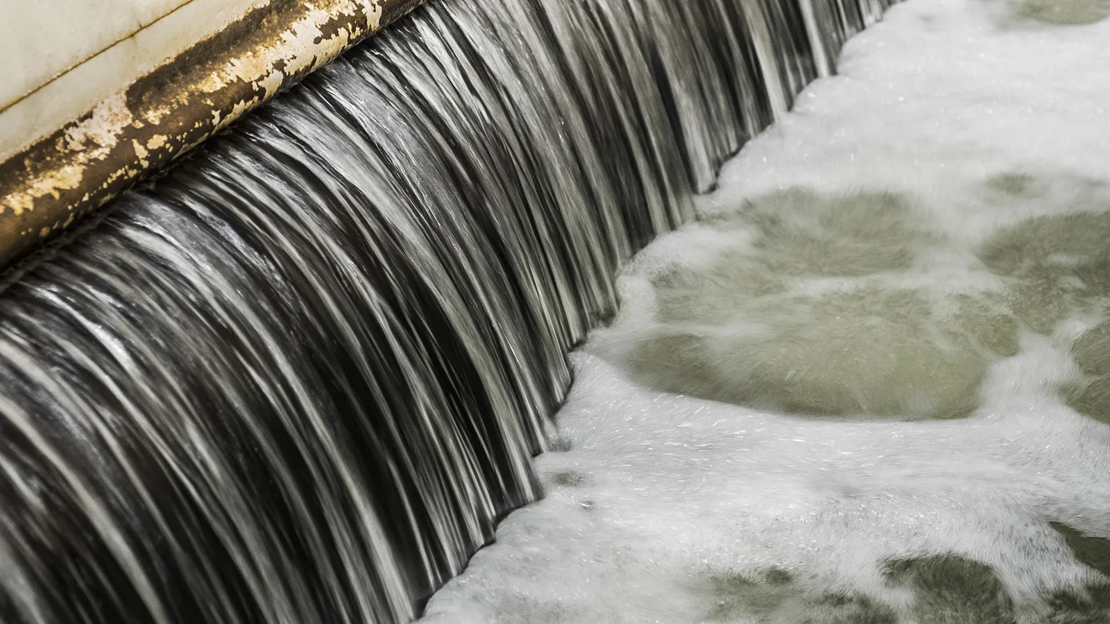 Vatten som rinner över en kant i en anläggning och bildar ett jämnt vattenfall, med skummande, bubbligt vatten nedanför.