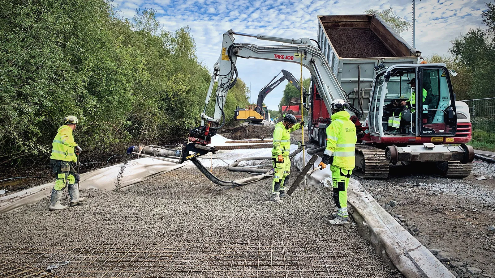Blåsläggning av Leca lättklinker på byggarbetsplats, där materialet appliceras via slang ovanpå armeringsnät med grävmaskin och arbetslag i arbete.
