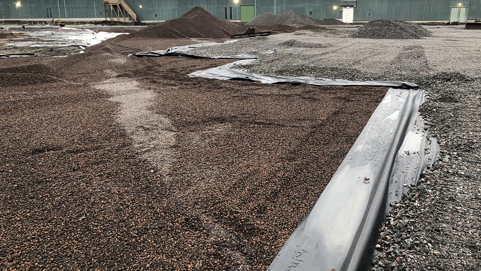 Yta med utlagd lättklinker ovanpå geotextil på en byggarbetsplats, där materialet formas i sektioner och omges av grus och upplag i bakgrunden.
