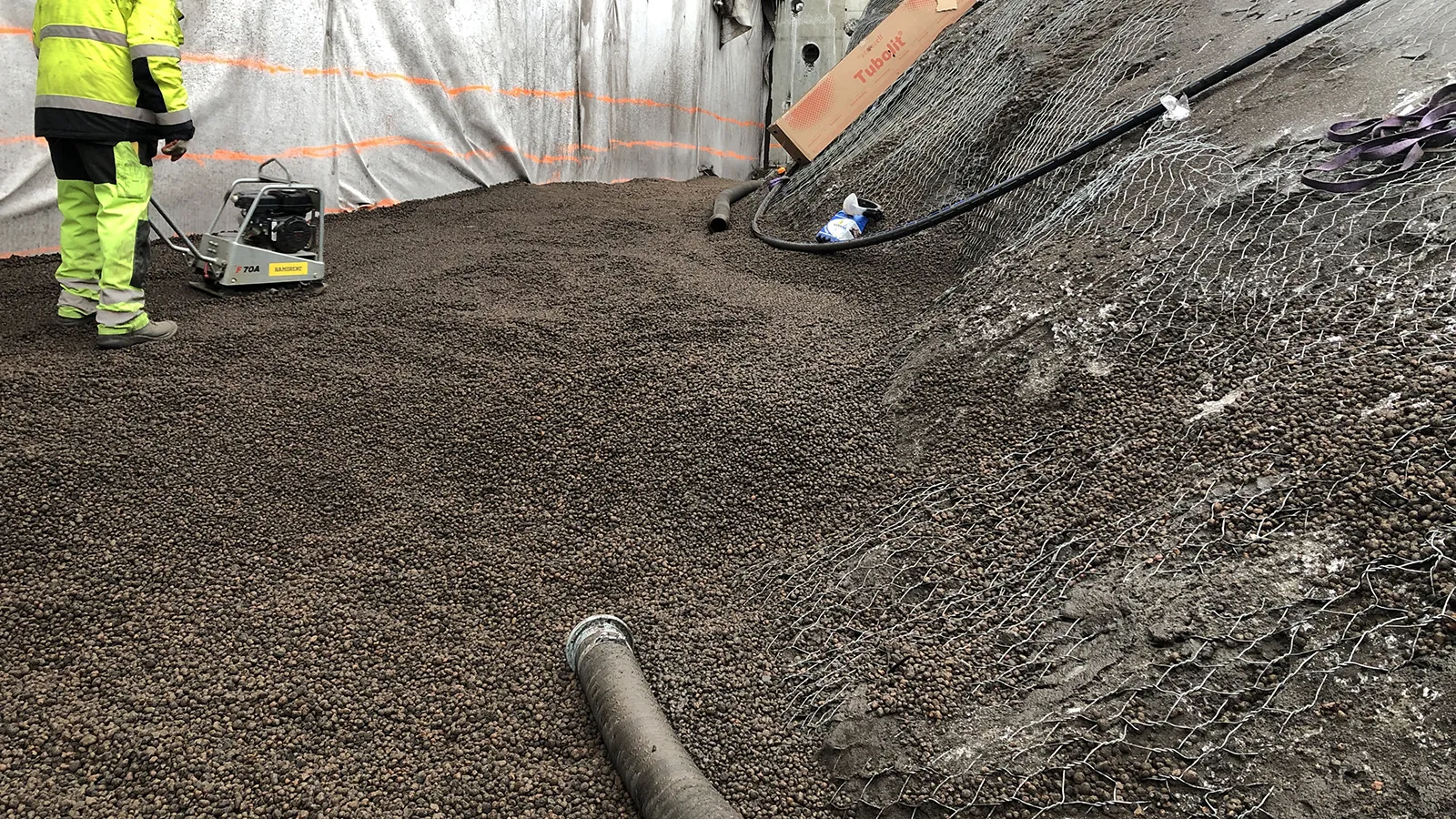 Installation av Leca lättklinker som lättfyllnad i slänt, där materialet packas med vibroplatta intill geonät och rörgenomföring.