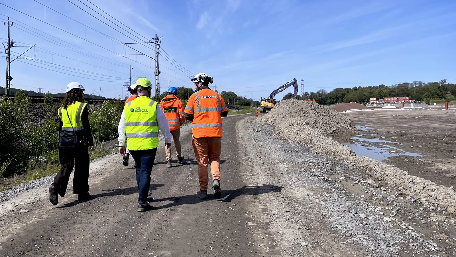 Fyra personer i varselkläder och hjälmar går längs en grusväg på en byggarbetsplats intill en järnväg. I bakgrunden syns en grävmaskin, jordhögar och ett öppet arbetsområde under klar himmel.