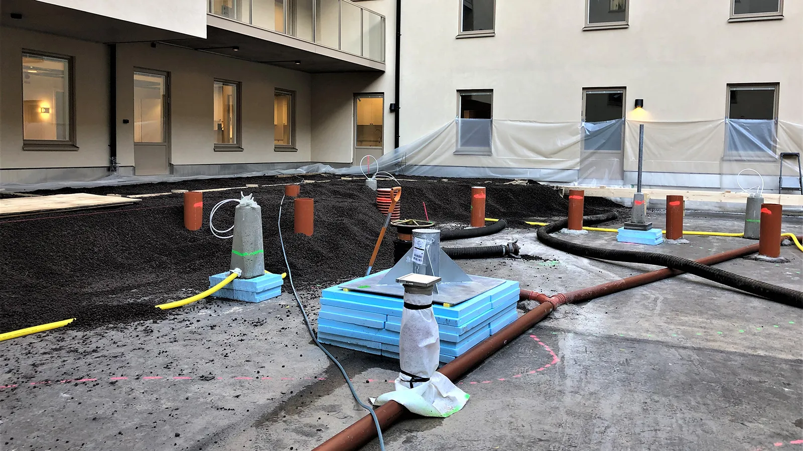 Innergård under byggnation med installationer för mark- och VA-arbete, där rör, kablar och isolerade fundament är utplacerade på en gjuten betongyta, omgiven av bostadsfasader.