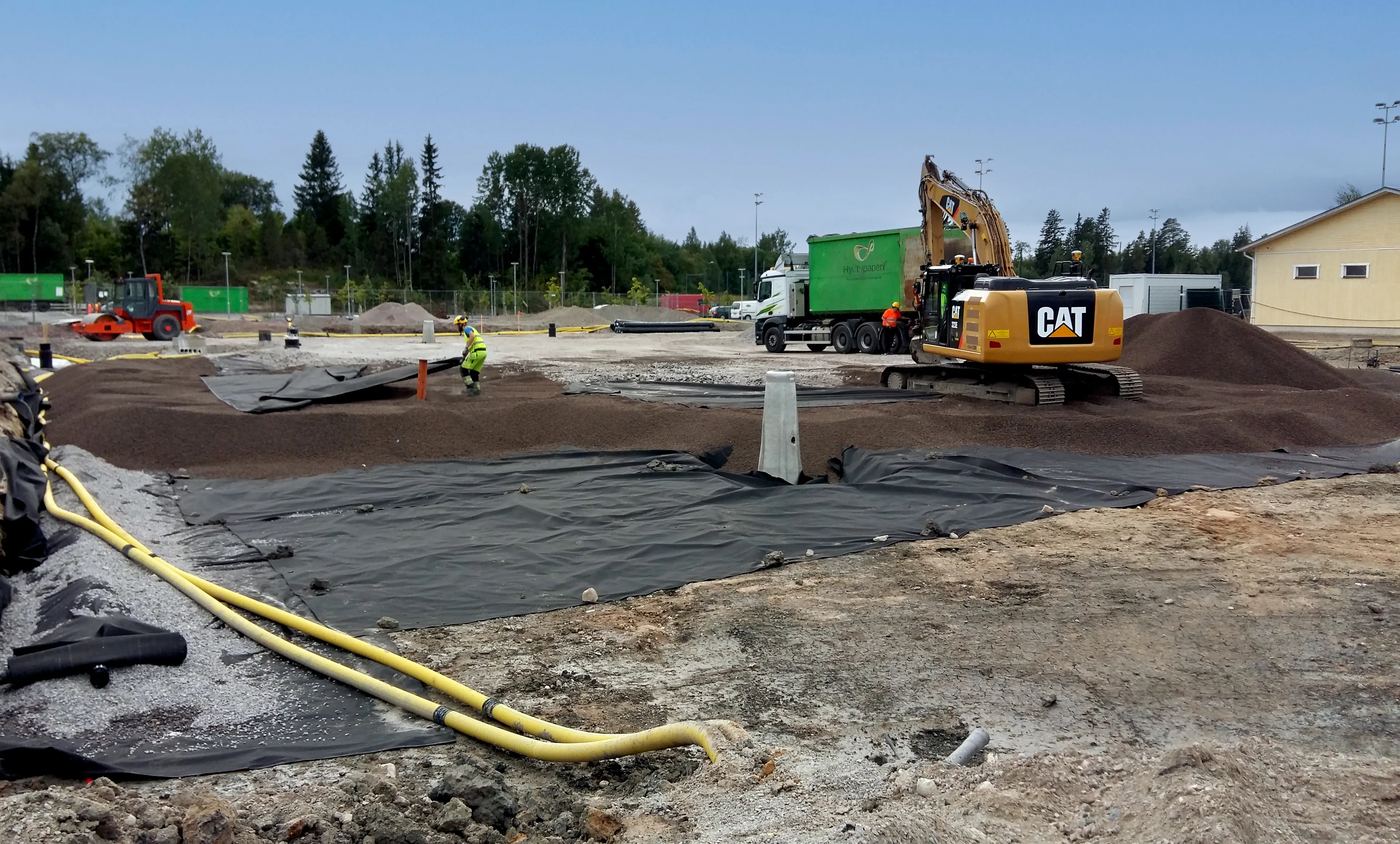 Byggarbetsplats där lättklinker läggs ut som lättfyllnad ovanpå geotextil, med grävmaskin, lastbil och arbetare i arbete.