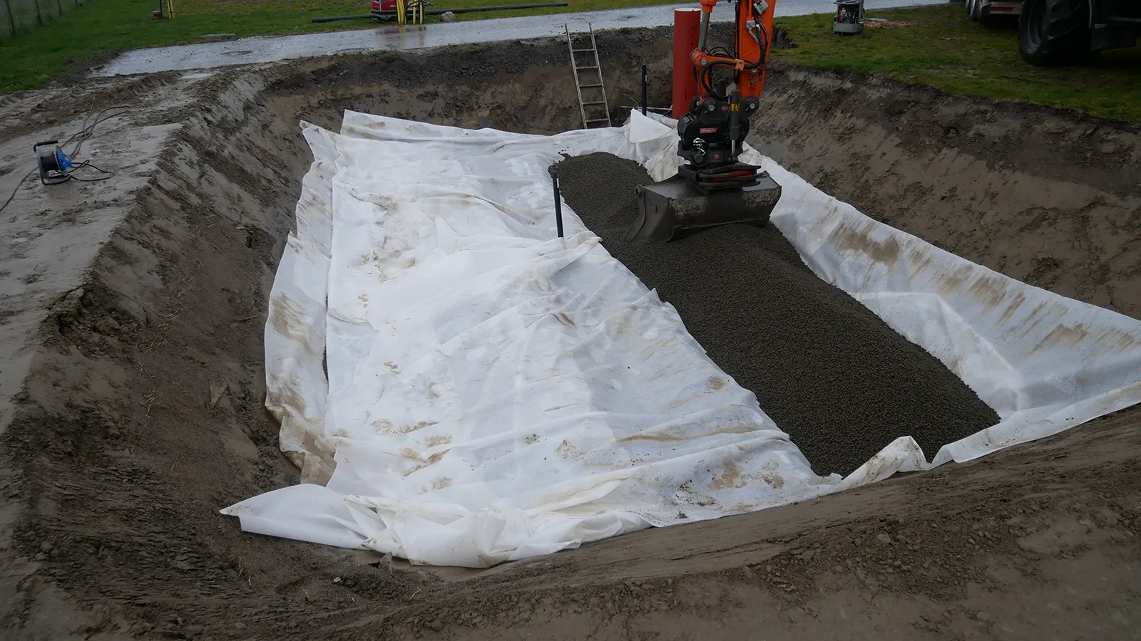 Schakt där Leca lättklinker läggs ut som lättfyllnad i geotextilklädd grop, med grävmaskin som fördelar materialet.