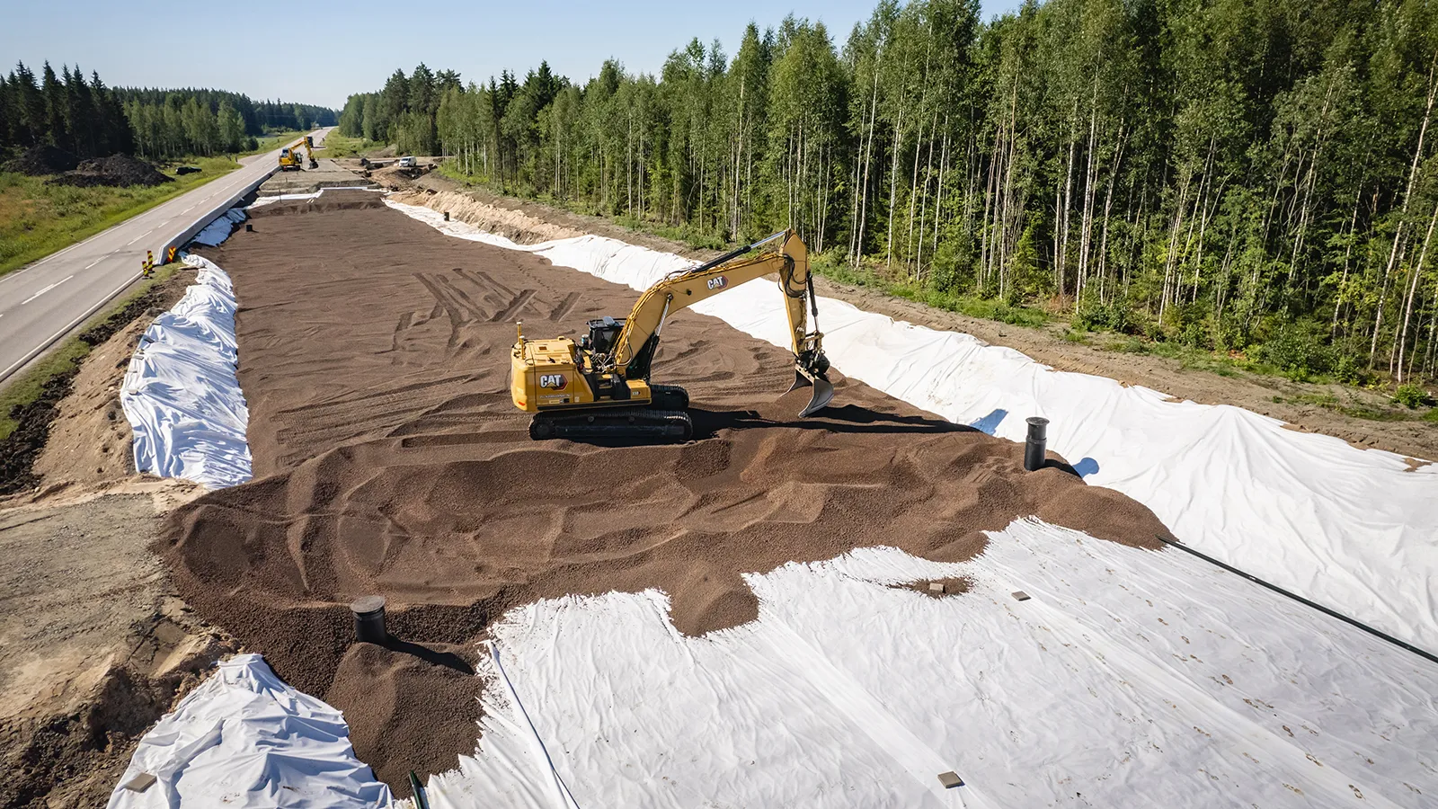 Vägbygge där lättklinker används som lättfyllnad ovanpå geotextil, med grävmaskin som sprider material längs vägsträcka intill skog.