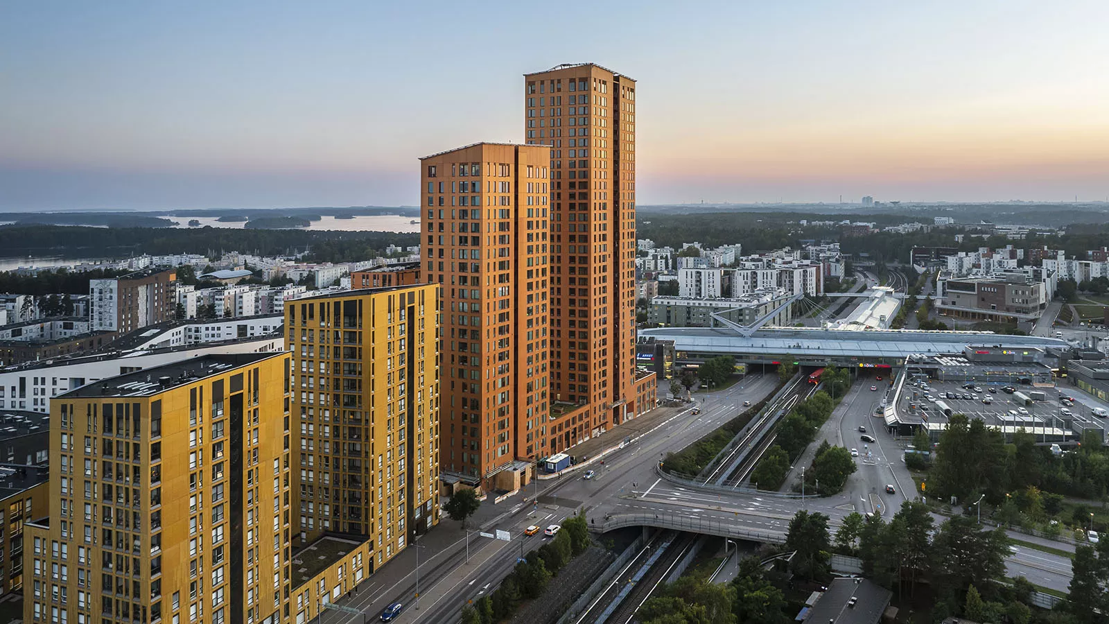 Flygfoto över ett stadslandskap med två höga orange byggnader, gröna områden och en sjö i bakgrunden.