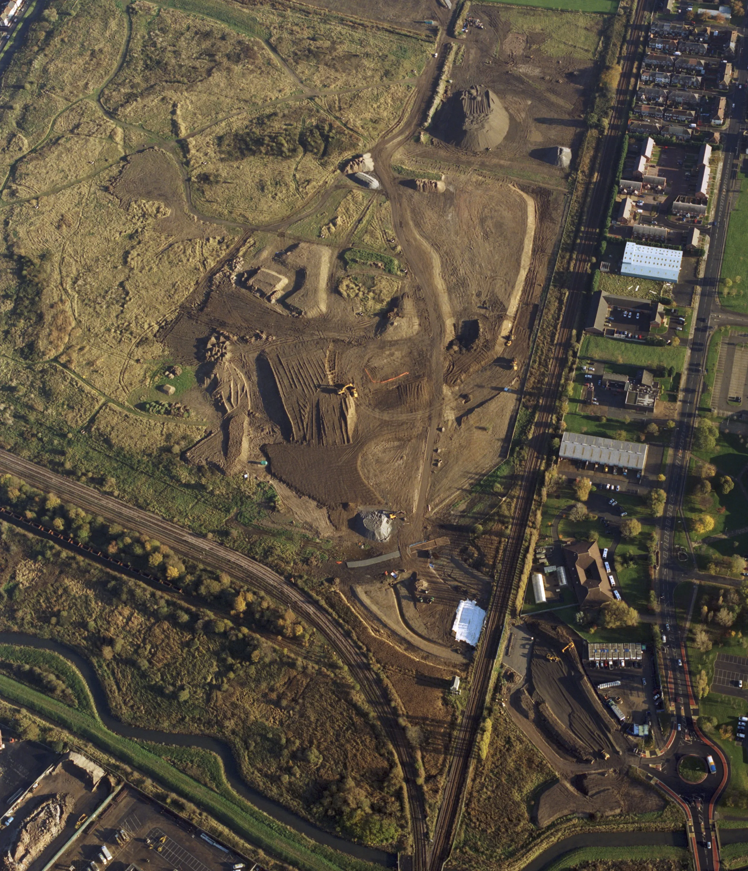 Flygbild över ett större mark- och infrastrukturprojekt där schaktning, arbetsvägar och upplag av material syns intill järnväg och bebyggelse.