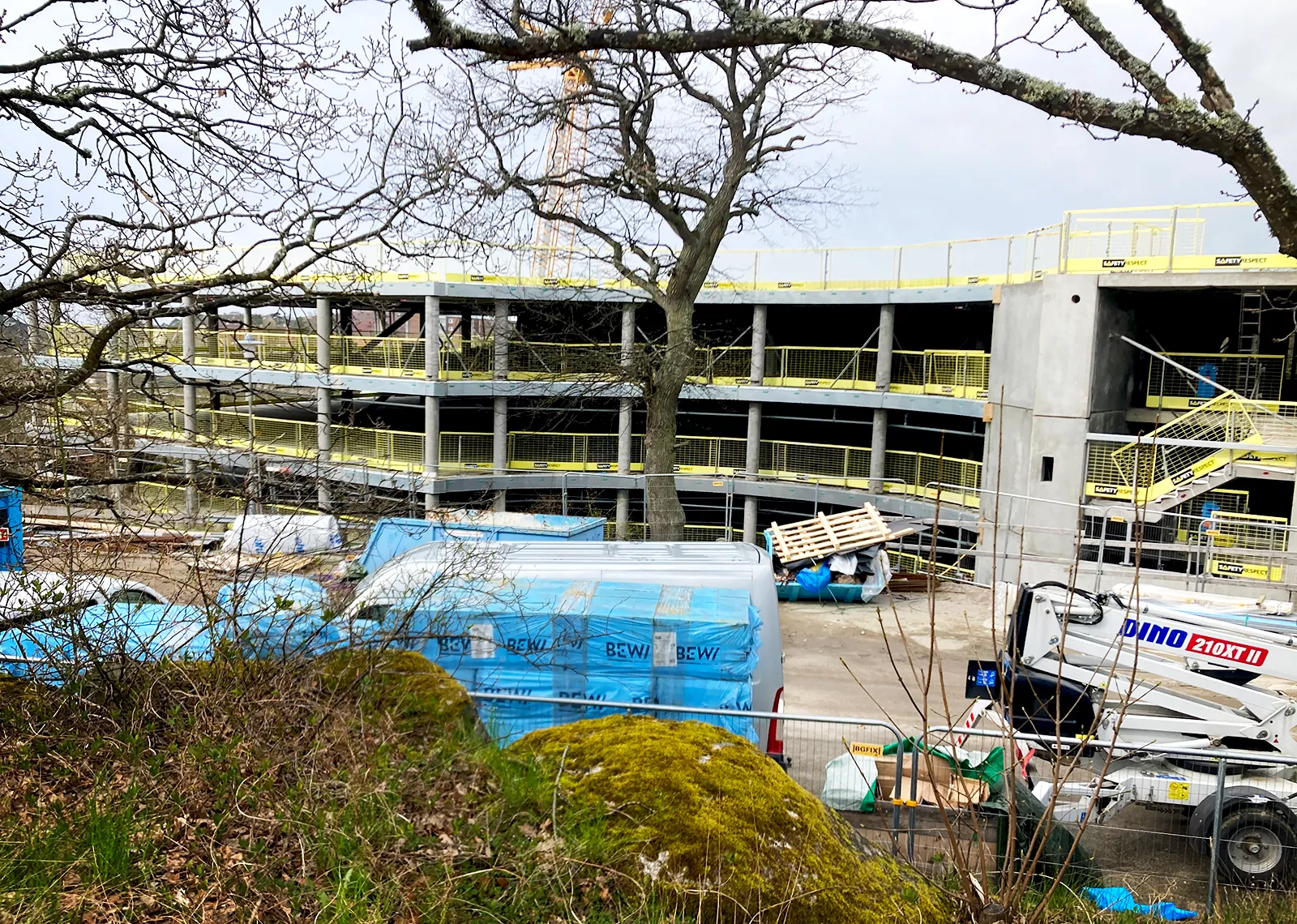  Översiktsbild över en byggarbetsplats med en betongkonstruktion i flera våningar. Förgrunden visar lastade byggmaterial och mossbeklädda stenar, med trädgrenar som ramar in motivet.