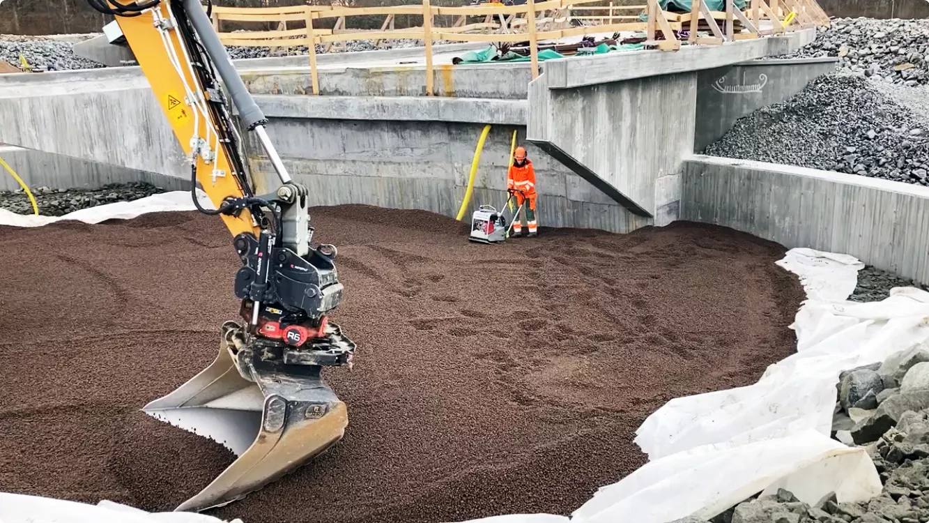 En grävmaskin arbetar med att jämna ut Leca Lättklinker i en grop, omgiven av betongväggar och geotextilduk. En arbetare i orange skyddsutrustning syns i bakgrunden med en markvibrator.