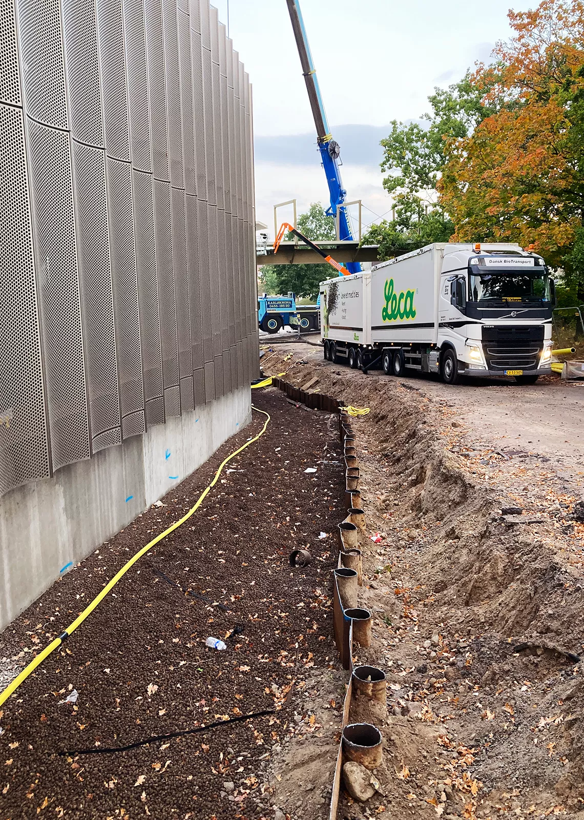 Leca-lastbil på byggarbetsplats med pågående fyllningsarbete längs en husfasad. Lättklinker har blåsts ut och fyller området mellan byggnaden och spontad jordkant.