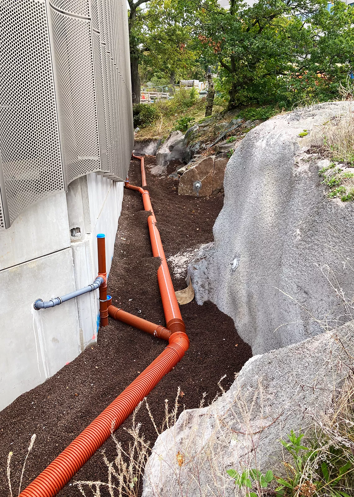 Dräneringsrör och avloppsledningar i orange plast installerade längs en byggnadsgrund, omgivna av lättklinkerfyllning och bergväggar i naturligt landskap.