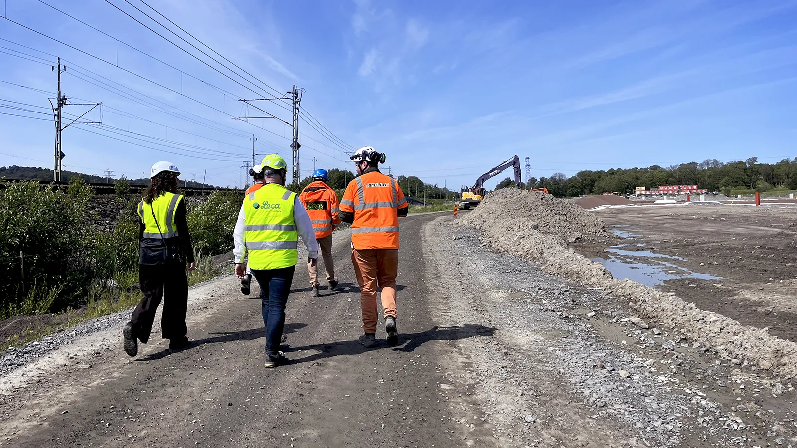 Platsbesök på infrastrukturprojekt med projektteam i varselkläder längs byggväg, intill järnväg och pågående markarbeten