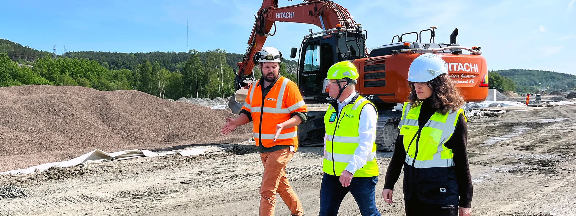 Tre personer i skyddsutrustning går bredvid en grävmaskin och högar med Leca lättklinker på en byggarbetsplats.