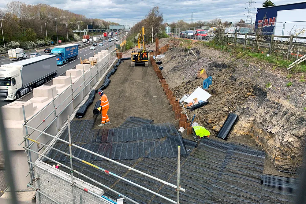 Anläggningsarbete vid en motorväg där geonät installeras i ett schakt för att stabilisera marken. En arbetare i orange varselkläder inspekterar ytan, medan en grävmaskin står redo längre bort. Tung trafik rullar förbi på motorvägen intill, skyddad av en tillfällig betongbarriär.