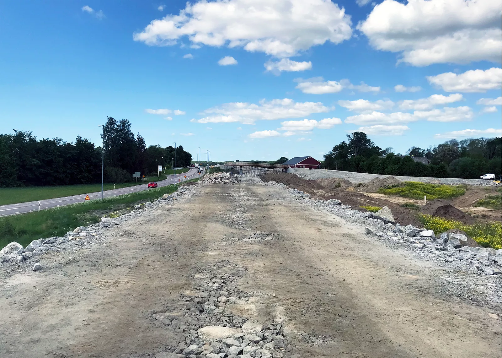 En stor grusyta under konstruktion med en väg som löper parallellt. Arbetet förbereder marken för vidare byggnation, med grönska och några träd längs vägen.