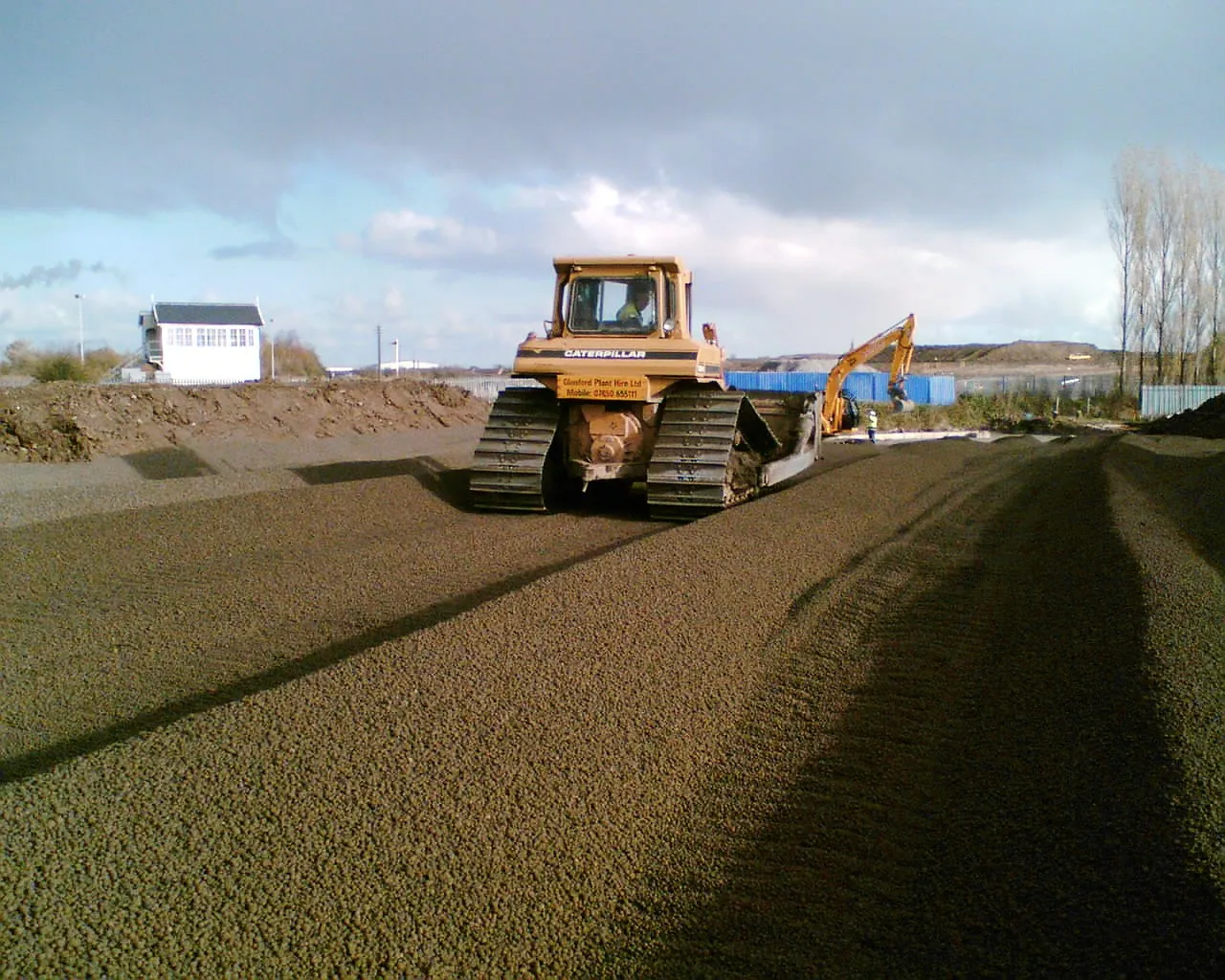 Bandtraktor jämnar ut Leca lättklinker på en byggarbetsplats, med grävmaskin och materialupplag i bakgrunden.