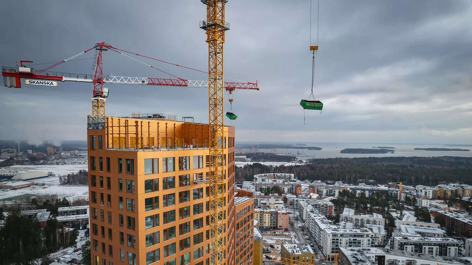 Närbild av en kran som lyfter en grön container med Leca-material över en hög byggnad i en urban miljö.