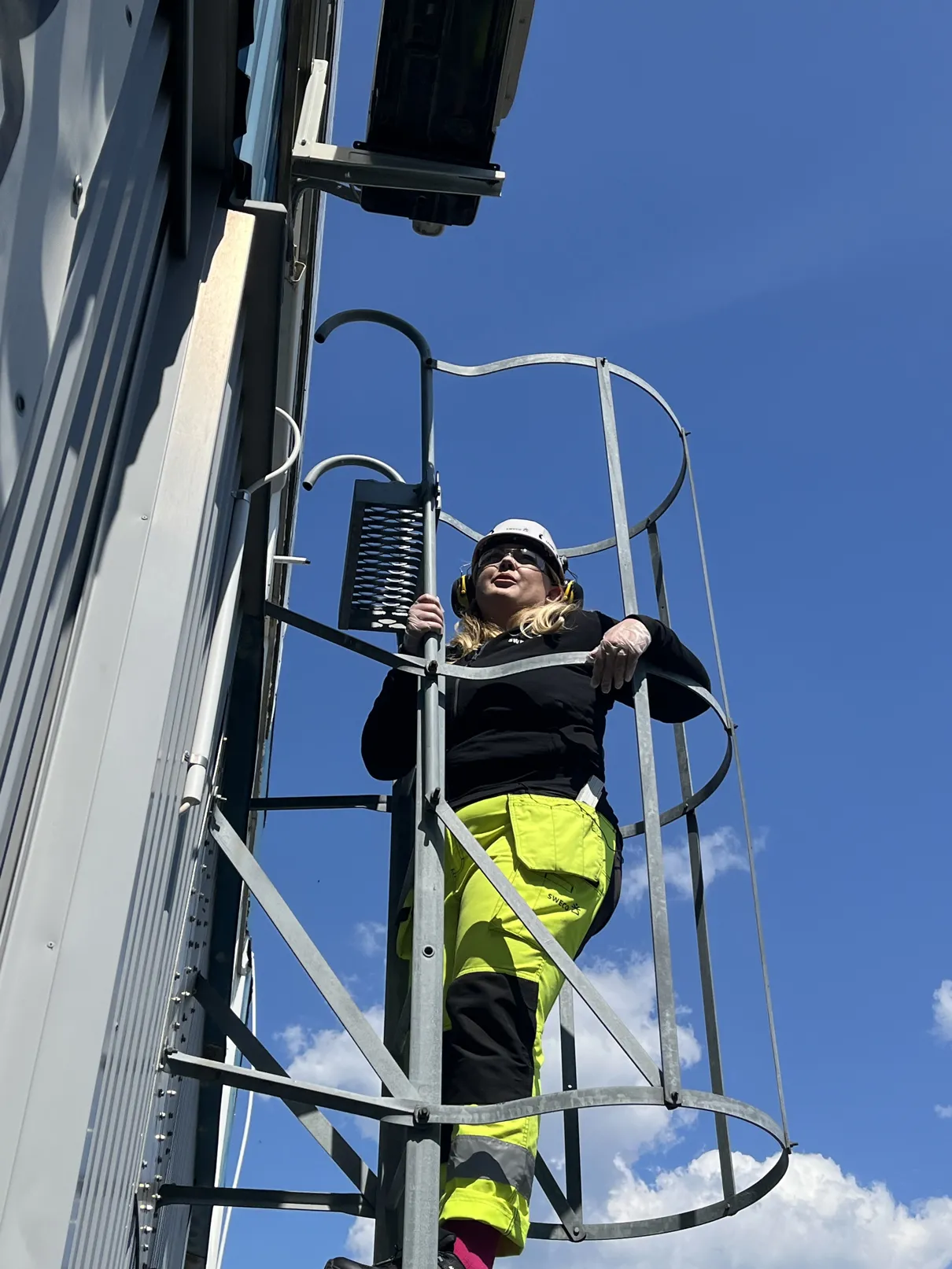 Kvinna i skyddskläder och hjälm klättrar uppför en stege på utsidan av en byggnad mot blå himmel.