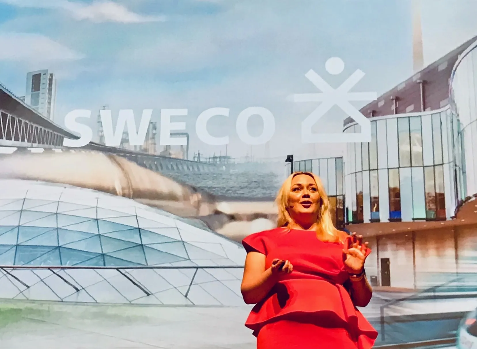 Amanda Borneke står på scen i röd klänning framför en stor bakgrund med Sweco-logotyp och stadsillustration.